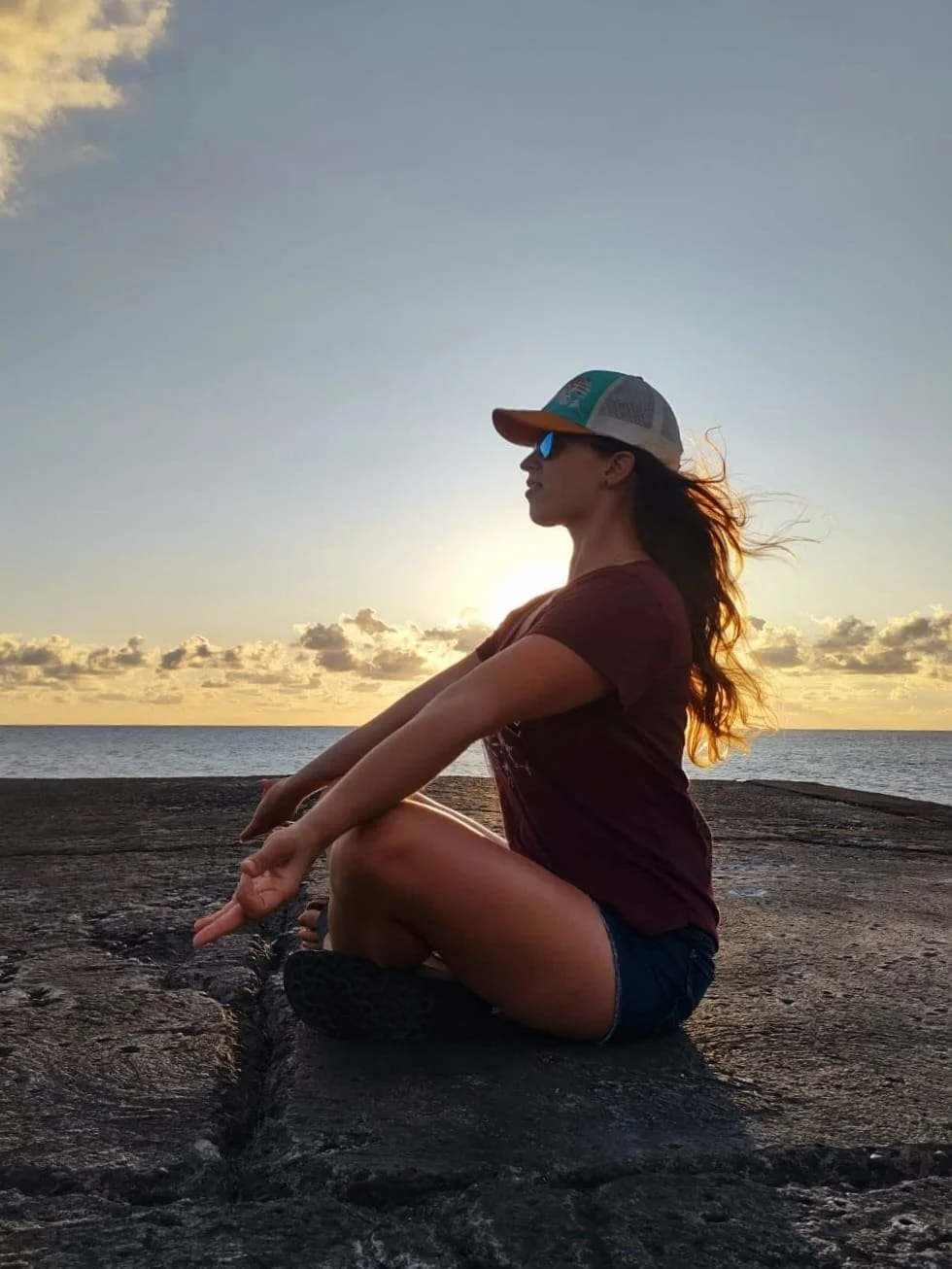 Une femme assise en méditation sur une roche au bord de la mer lors du coucher du soleil, portant une casquette, des lunettes de soleil, un t-shirt et un short denim.