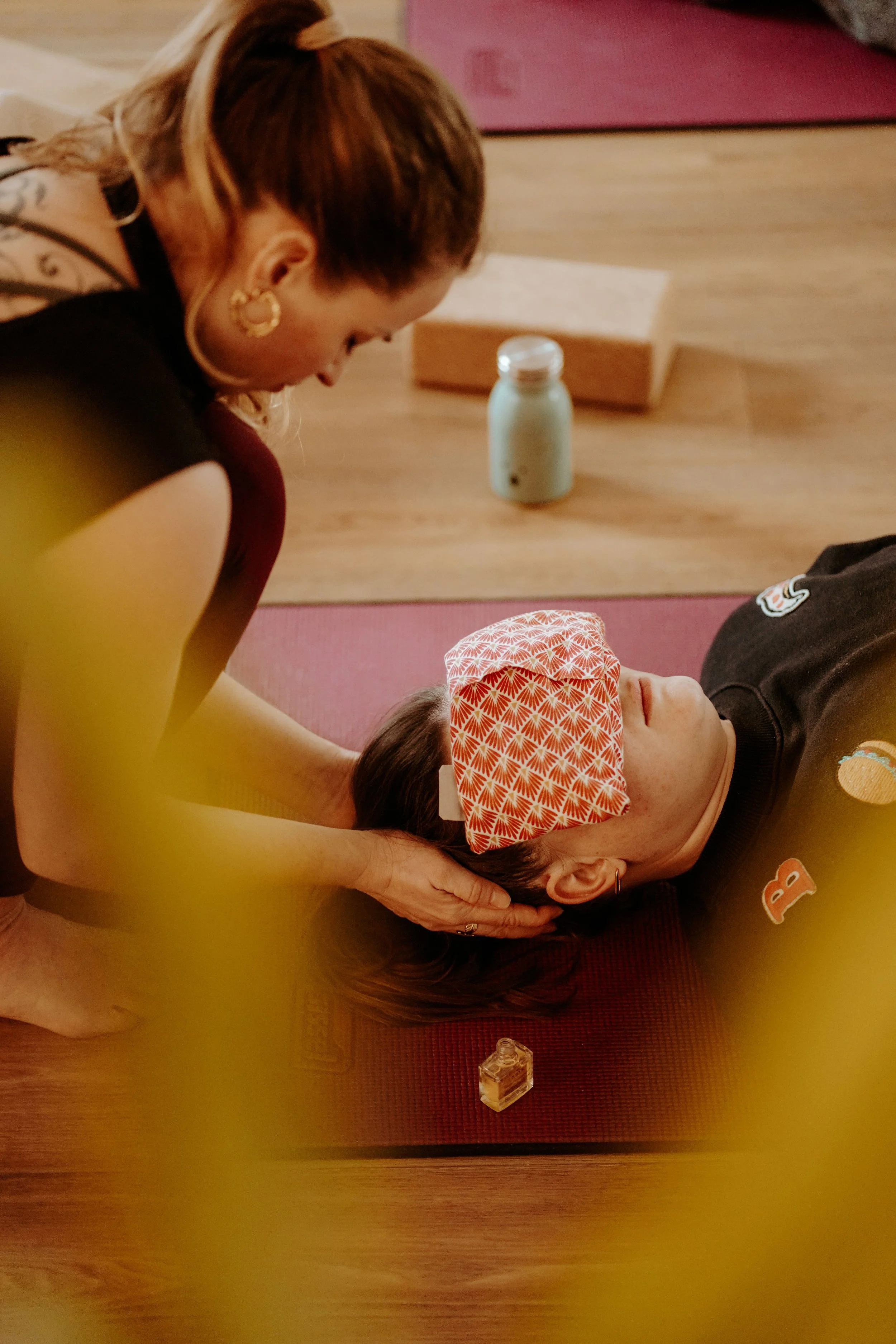 Une femme allongée sur un tapis de yoga avec un bandeau sur les yeux, pendant qu'une autre personne, une femme avec des boucles d'oreilles et un tatouage, lui soutient la tête avec les mains. On voit des blocs de yoga et un thermos derrière eux.
