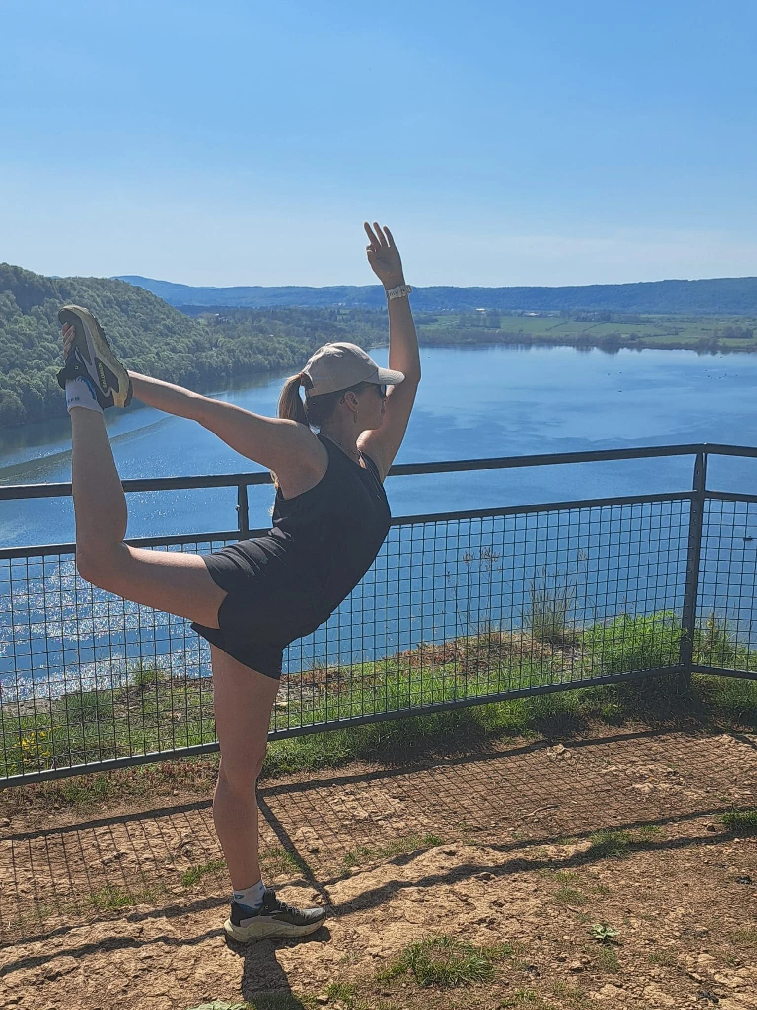 Une femme faisant une posture de yoga, debout en équilibre sur une jambe, tenant l'autre jambe derrière elle avec une main et levant l'autre bras vers le ciel, avec un paysage de rivière et de collines en arrière-plan.