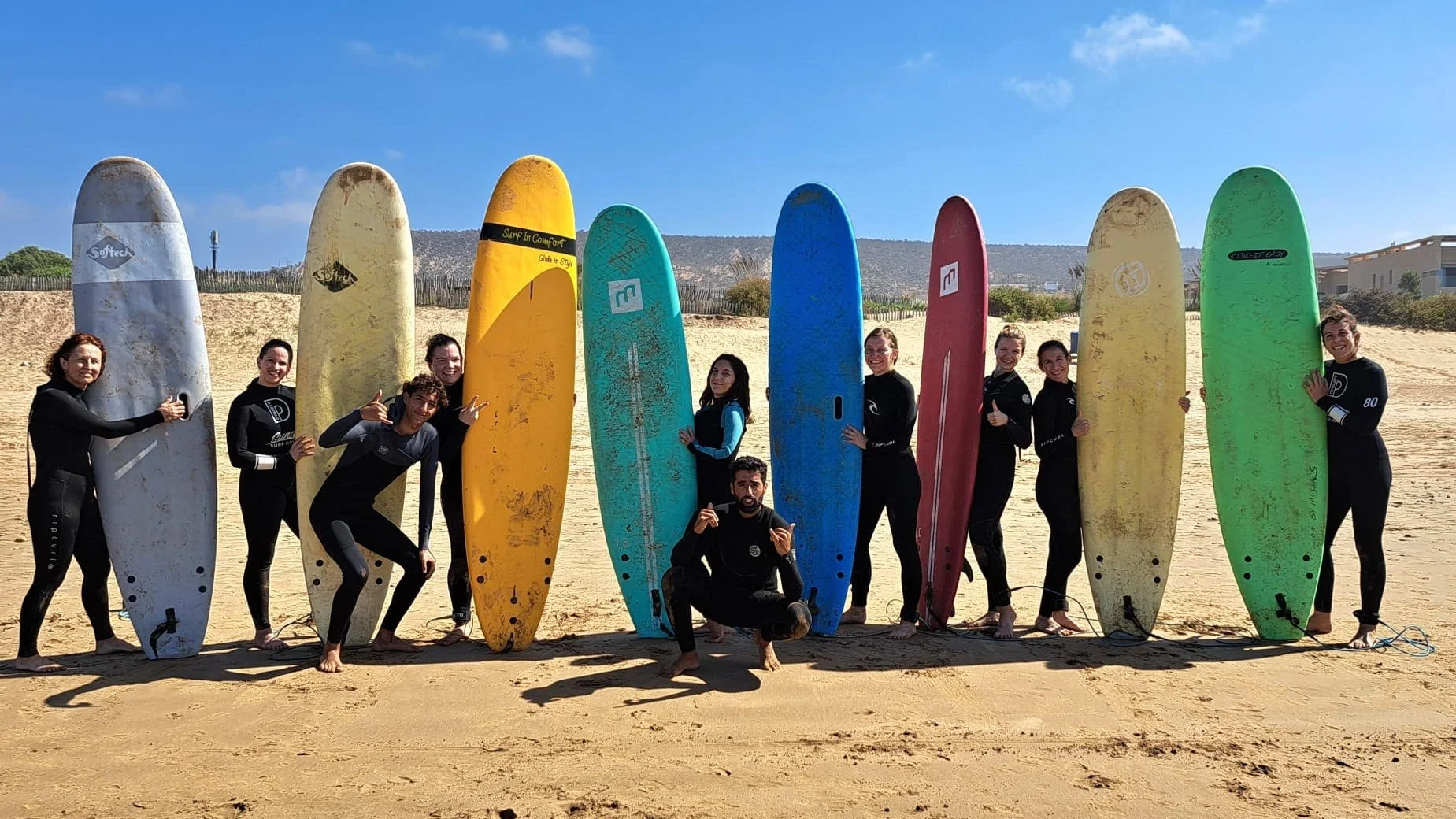 Groupe de personnes pendant un séjours surf et yoga en combinaison de surf, debout sur la plage, tenant des planches de surf colorées, avec un ciel ensoleillé en arrière-plan