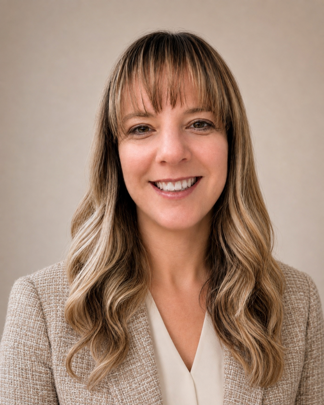 A woman with light brown, wavy hair, smiling, wearing a beige blazer and white blouse, standing against a plain neutral background.