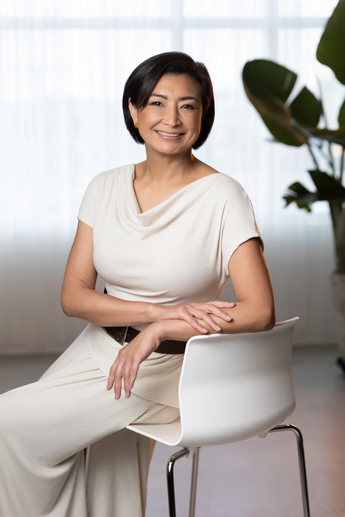 A woman with short black hair, wearing earrings and a light-colored dress, sitting on a white chair with X-shaped legs, smiling at the camera. There is a large window with sheer curtains and a plant with broad leaves in the background.