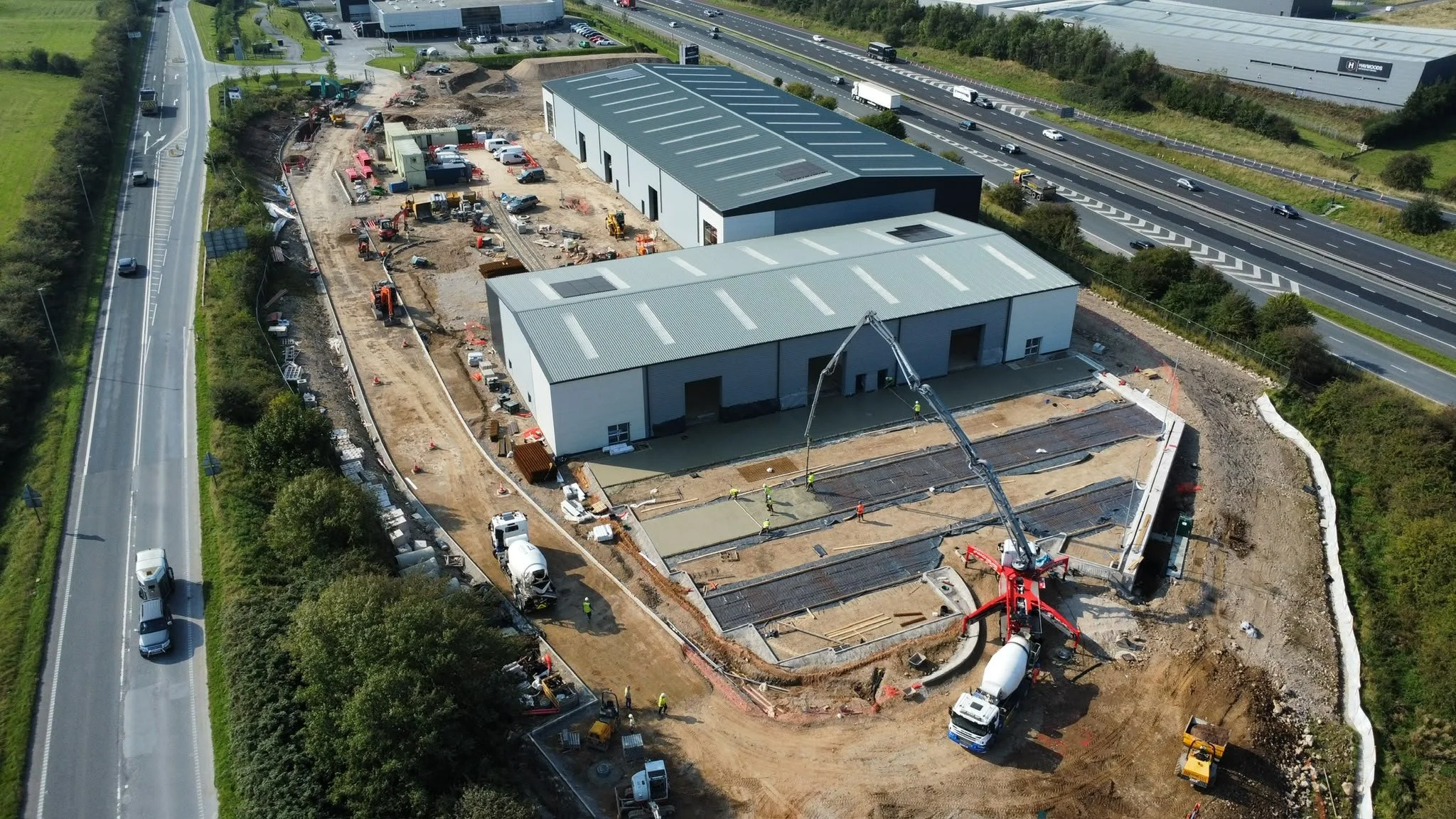An aerial view of a construction site next to a highway. Several buildings are under construction, with workers and building materials visible. Heavy machinery such as concrete mixers and cranes are actively working on the site.