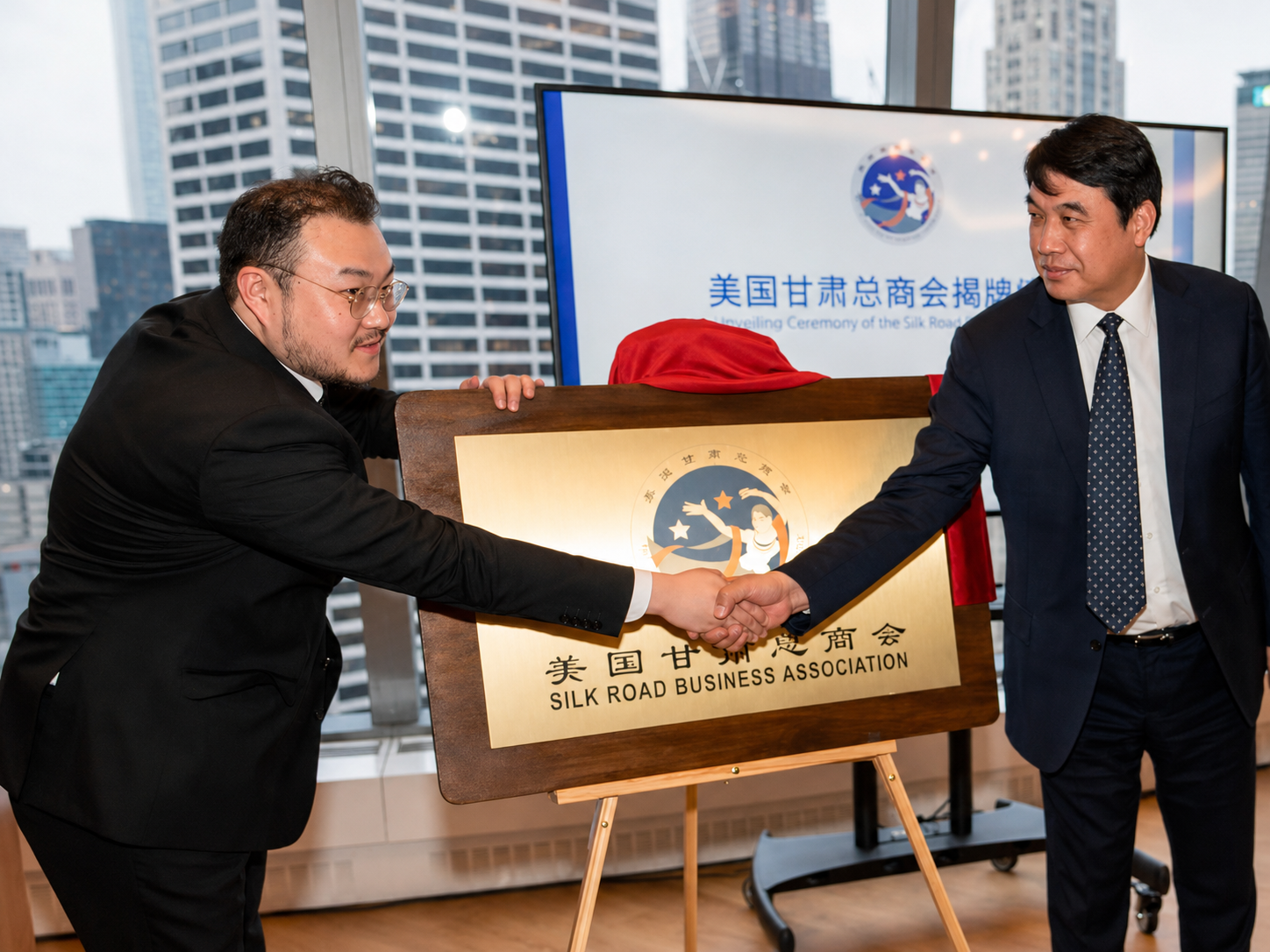 Two men in business suits shake hands during an unveiling ceremony for the Silk Road Business Association in a high-rise office with city buildings in the background.