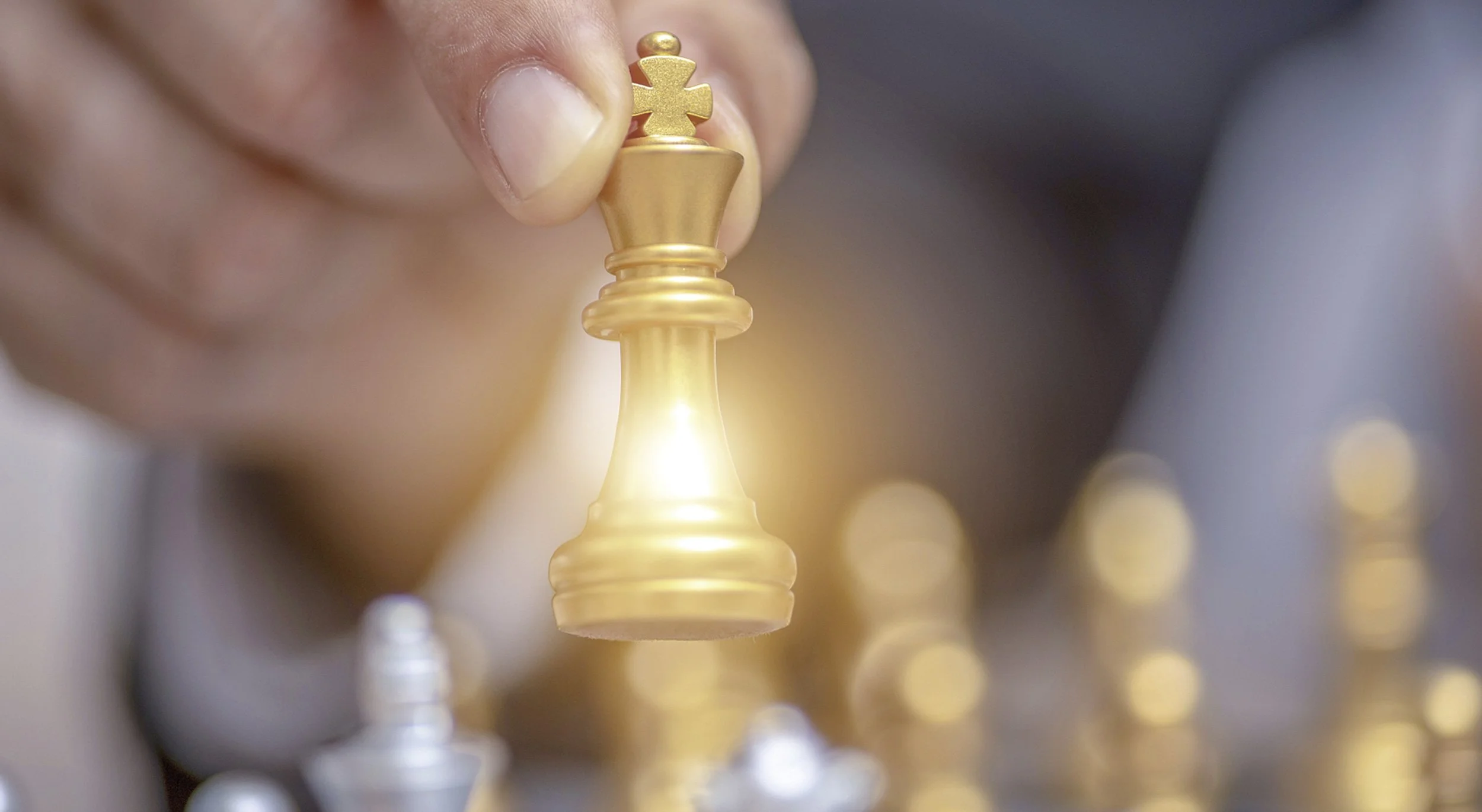 Close-up of a gold chess king piece on a chessboard, with other chess pieces blurred in the background.