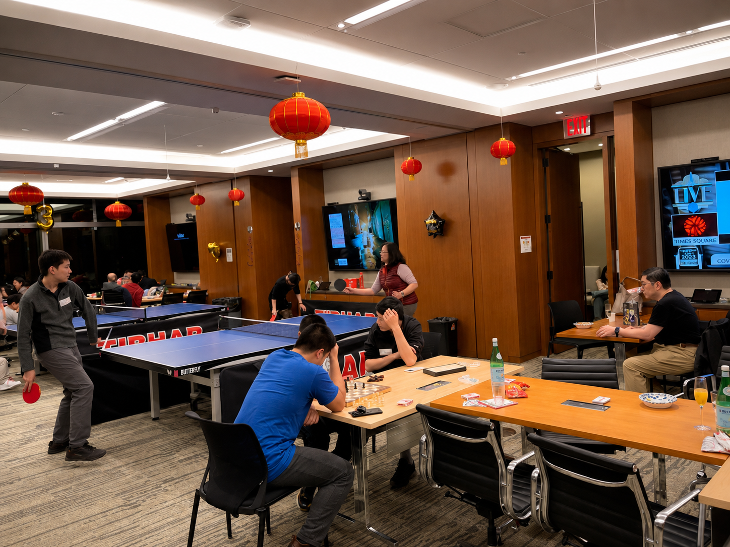 A lively indoor scene with people playing ping pong, sitting at tables, and watching a chess game. The room is decorated with red lanterns hanging from the ceiling, and several television screens and a large digital display are mounted on the walls. There are snacks and drinks on the tables, and some people are engaged in conversations and activities.