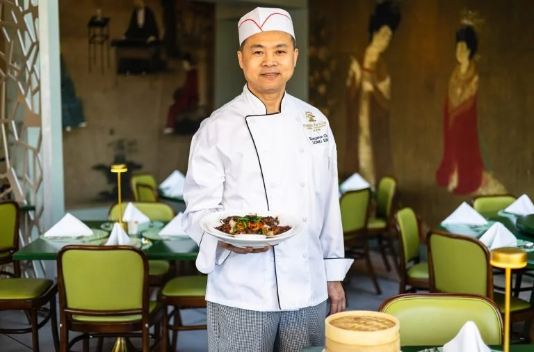 A chef in a white uniform and hat holding a plate of food in a restaurant with green chairs and elegant table settings, with traditional Asian artwork on the wall behind him.