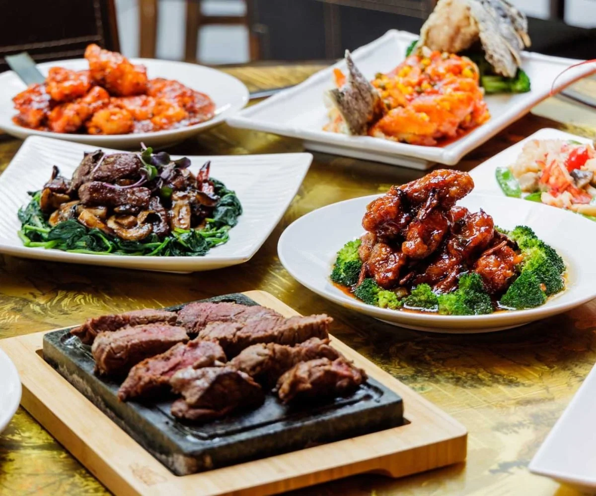Assorted Chinese dishes on plates and a wooden serving board, including sliced beef, stir-fried vegetables, sweet and sour chicken, and shredded crab with vegetables.