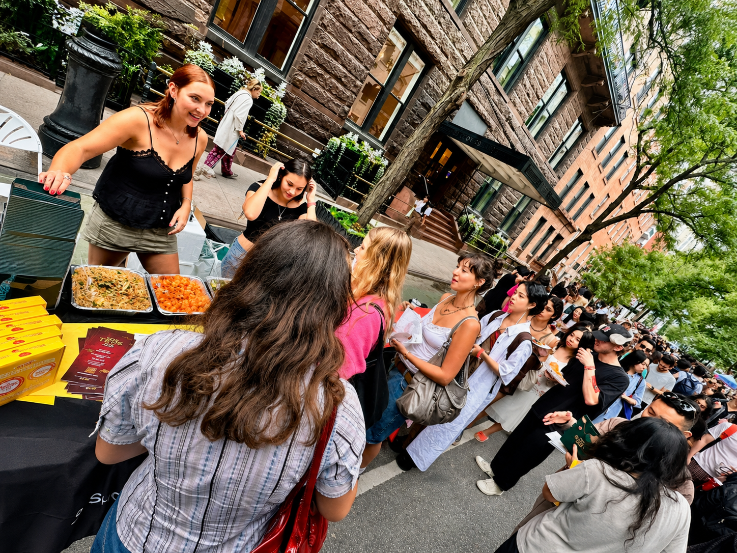 People lined up at a food vendor on a city street, with two women selling food and several customers waiting to buy.