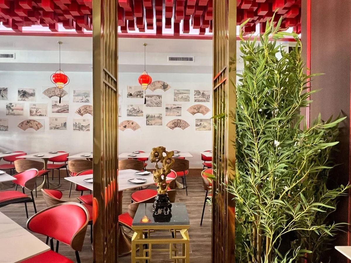 Interior of an Asian restaurant with red and wooden chairs, wall decorations of traditional Asian art, hanging red lanterns, and a decorative sculpture on a black and gold table near a green plant.