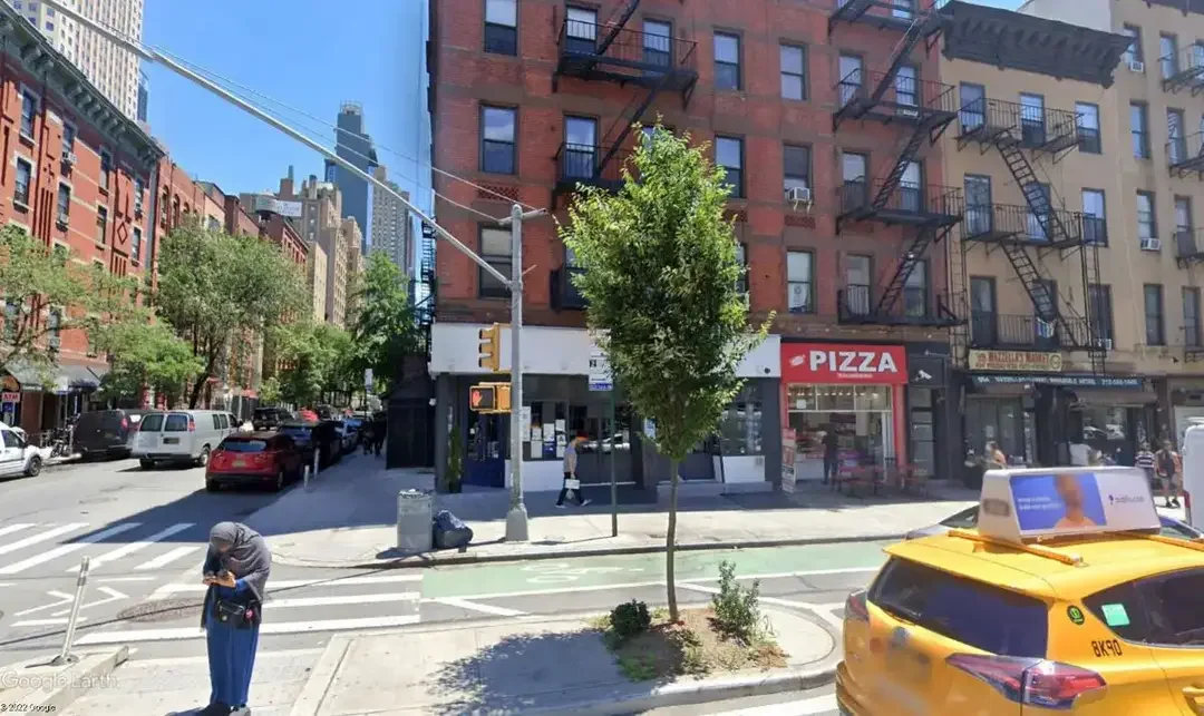 City street scene with a building that has a pizza restaurant and other shops, trees, and pedestrians, yellow taxi with a roof sign, and parked cars.