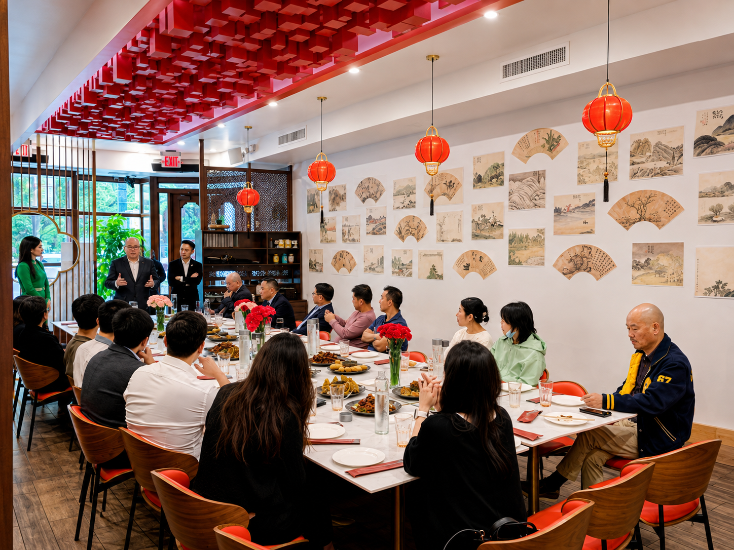 People attending a meeting or event in a restaurant with Asian decor, including red lanterns and traditional fan artwork on the walls, a long table with food, and speakers standing at the front.