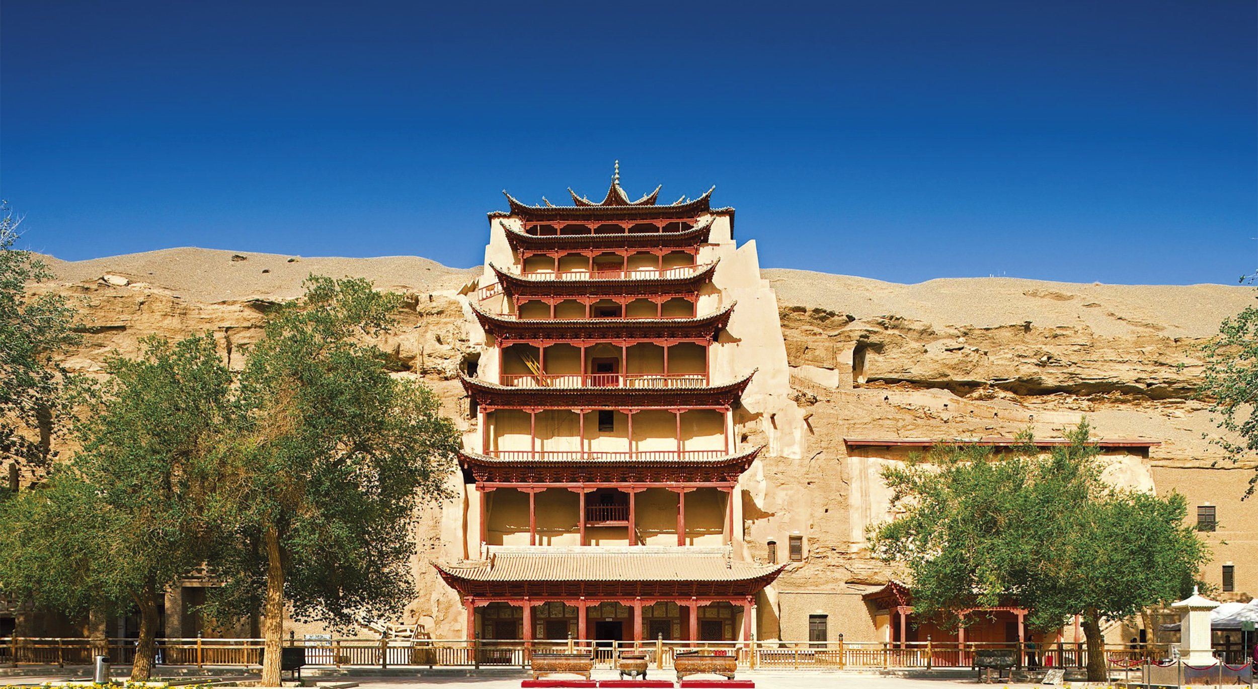 A multi-story Chinese-style building built into a rocky hillside, with layered, curved roofs and red wooden accents, flanked by green trees in front, under a bright blue sky.
