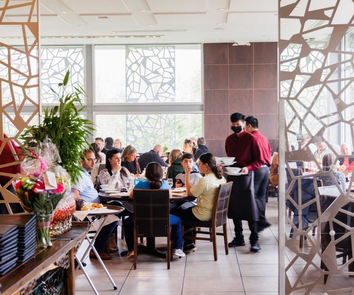 A busy restaurant interior with several diners seated at tables, and waitstaff serving food. Bright natural light filters through large decorative windows.