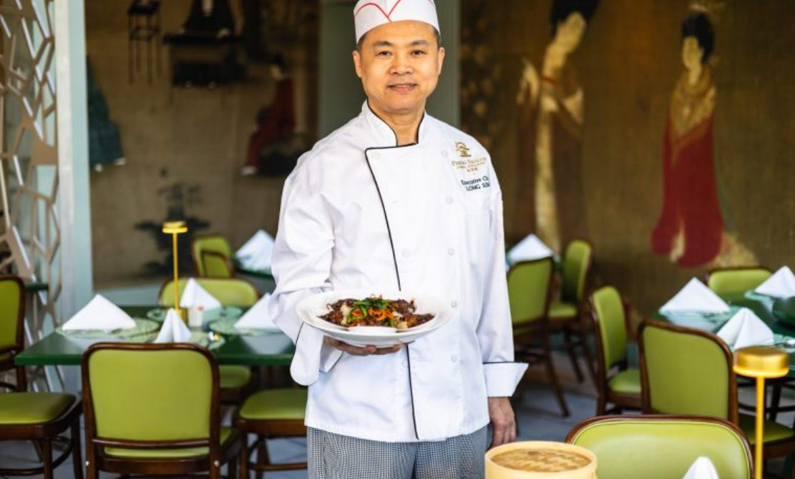 A chef in a white uniform holding a plate of food in a restaurant with green chairs and a mural on the wall.