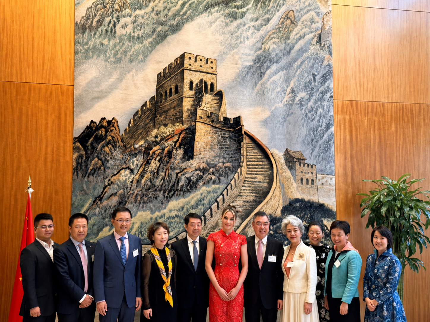 Group of ten people standing in front of a large artwork of the Great Wall of China, with wooden paneling on the sides and a flag on the left.