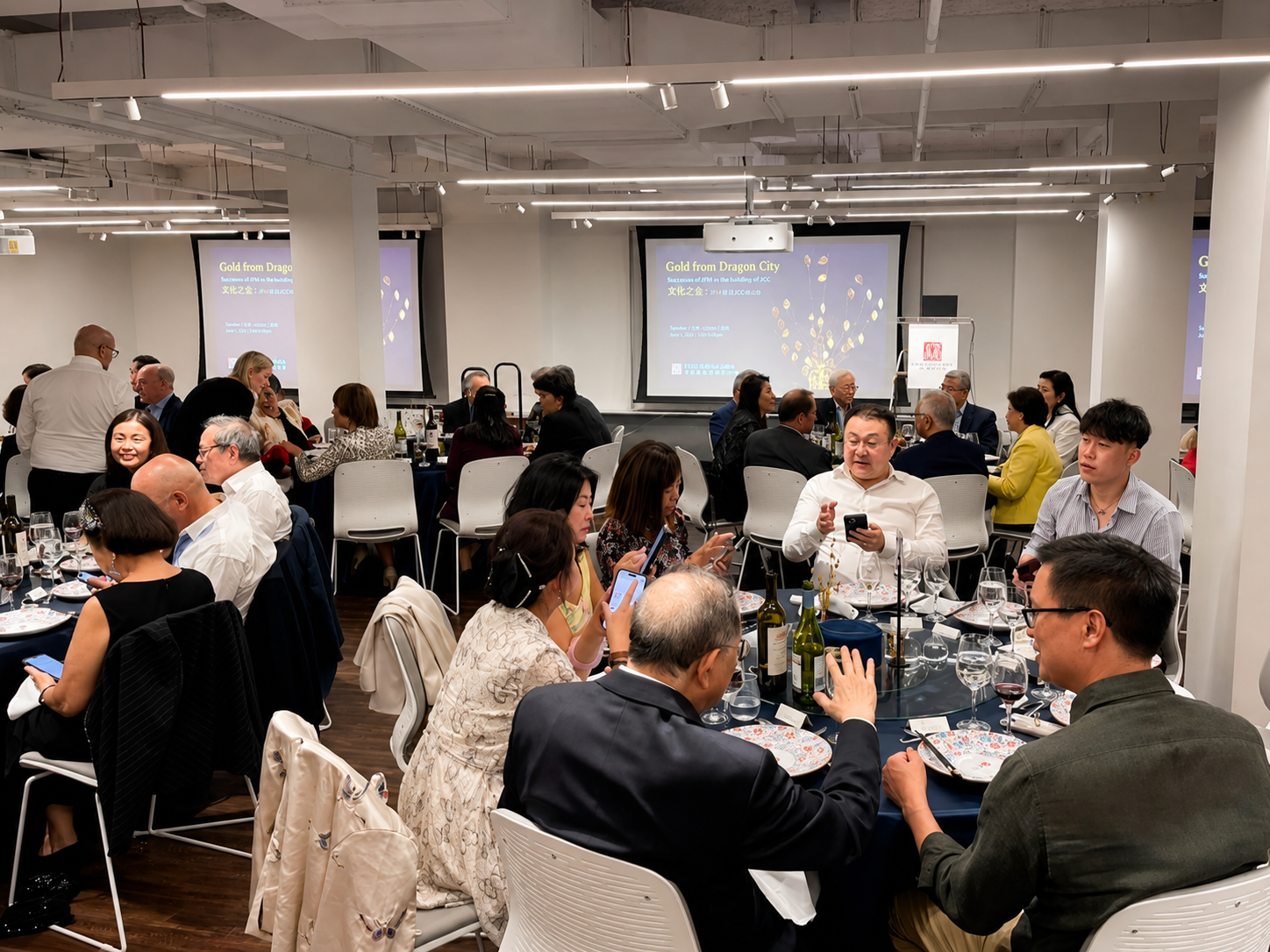 A formal dinner event with multiple round tables filled with people, some using their phones. There are three large screens at the front displaying a presentation titled 'Gold from Dragon City' with Chinese text, in a bright, modern conference room with white walls and ceiling lights.