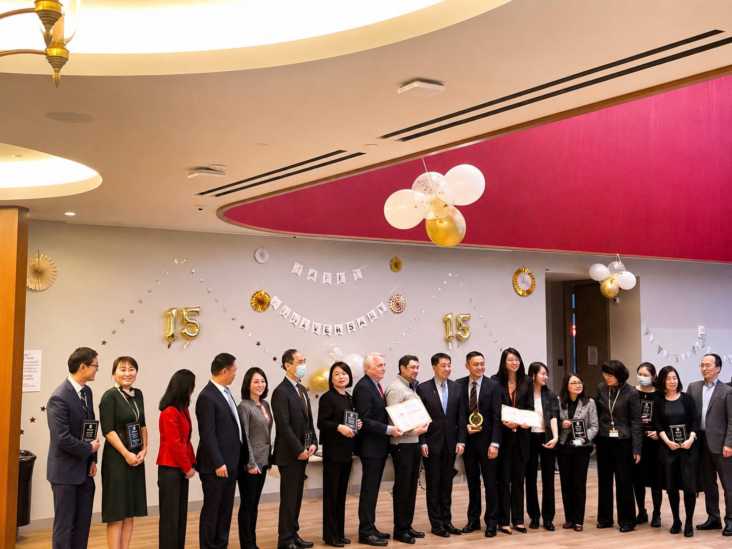 Group of people dressed in formal attire standing in a line during a celebration of::15th anniversary, with decorated wall including balloons, banners, and golden balloons displaying the number 15.