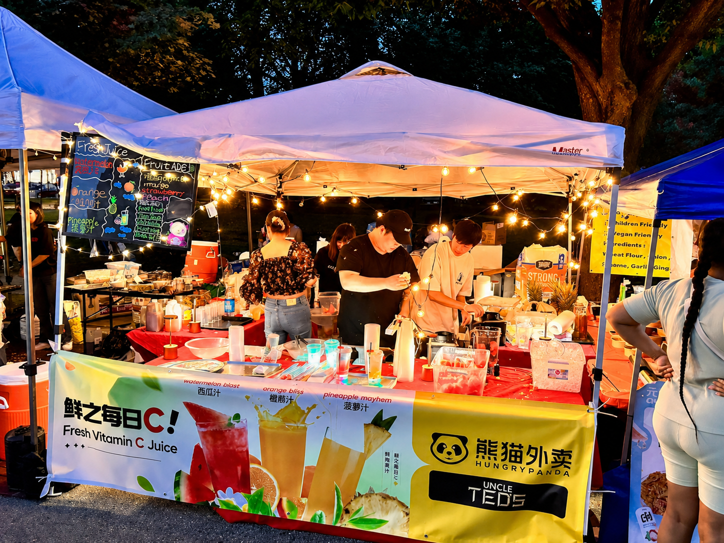 A lively outdoor juice stand at dusk with string lights, colorful banners, and people preparing drinks. The banner advertises fresh vitamin C juice with images of colorful fruit drinks and text in various languages.