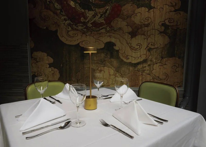 Elegant dining table set for four with white tablecloth, white folded napkins, wine glasses, water glasses, and silverware, illuminated by a small gold table lamp, with a decorative mural in the background.