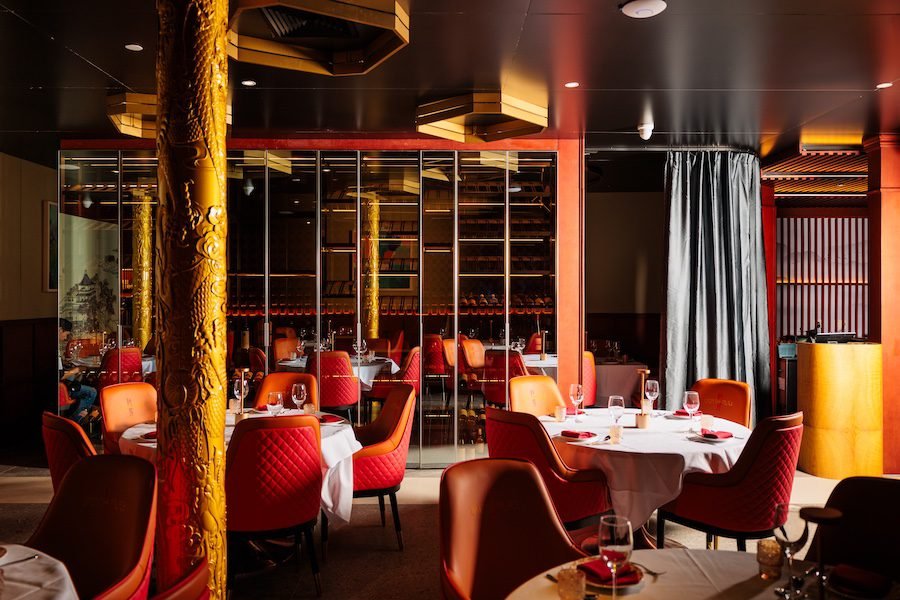 Elegant restaurant with round tables covered with white tablecloths, set with wine glasses, utensils, and napkins. Red and orange upholstered chairs surround the tables. The room features decorative golden columns, a mirrored wall, and dark ceiling panels. A gray curtain hangs beside a yellow curved partition.