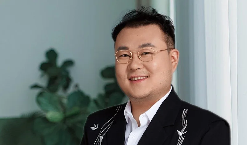 A smiling man in a black blazer with white embroidery, white shirt, and glasses, standing indoors in front of a window and green plants.