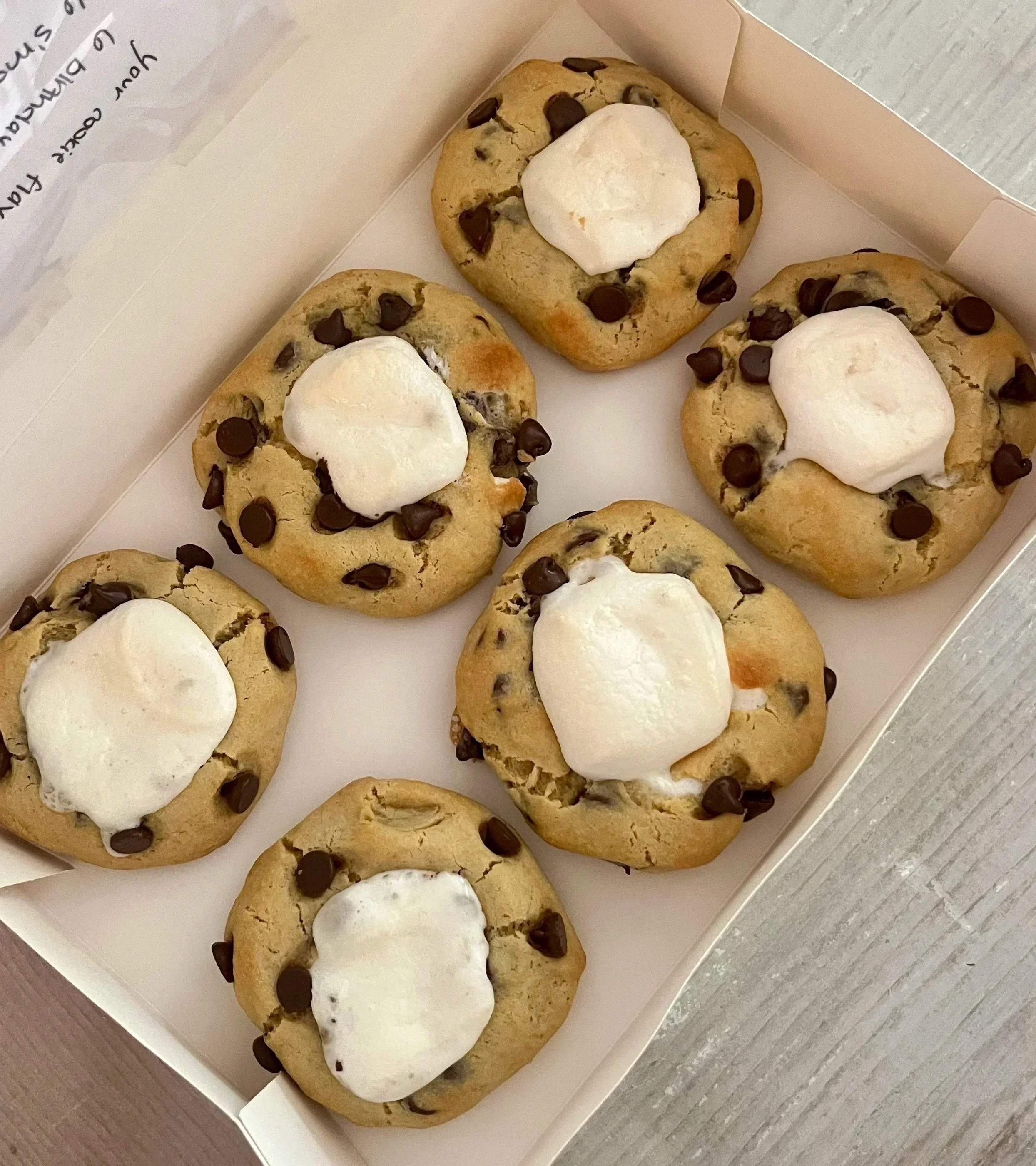 Close-up of five cookies with chocolate chips and a dollop of marshmallow, placed in a white box on a gray surface.