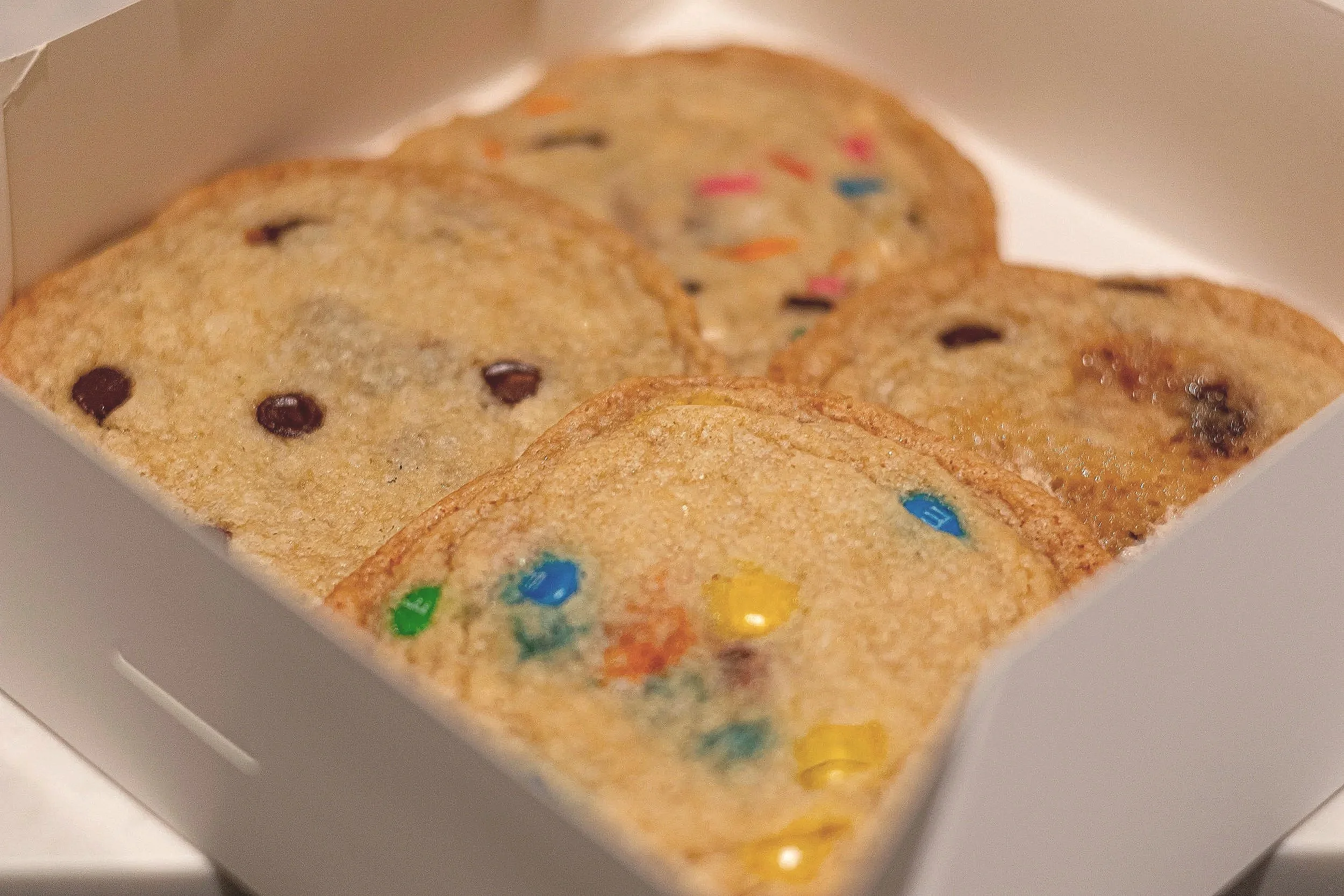 Close-up of a white box holding three decorated cookies with colorful sprinkles and chocolate chips.