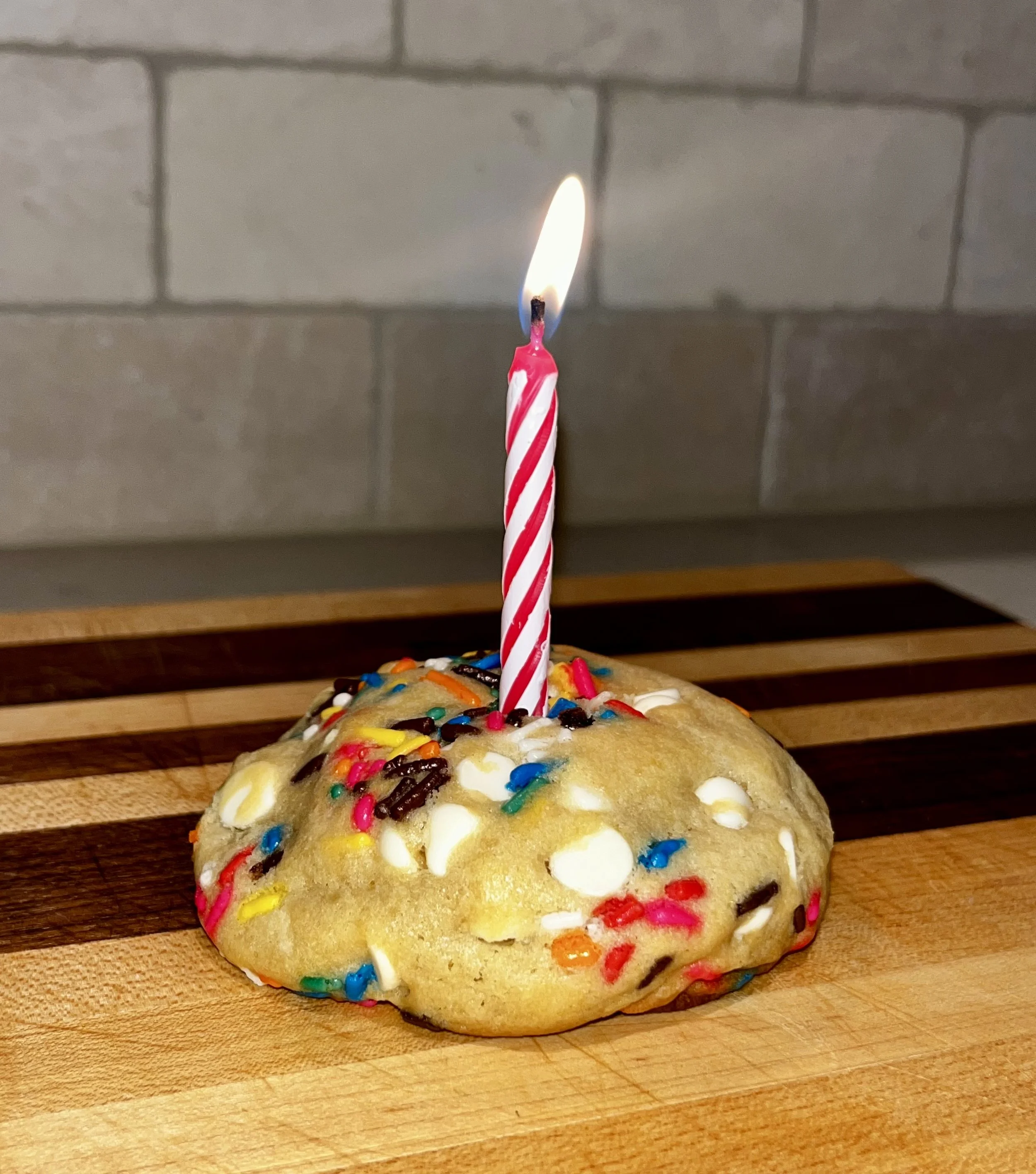 A birthday cake cookie decorated with colorful sprinkles and white chocolate chips, with a birthday candle on top. 