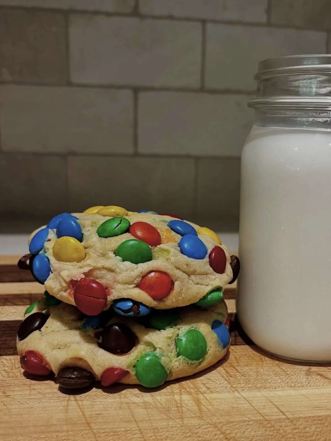 Two colorful candy cookies with M&M candies, placed on a wooden cutting board with a jar of milk beside them, in front of a brick wall.