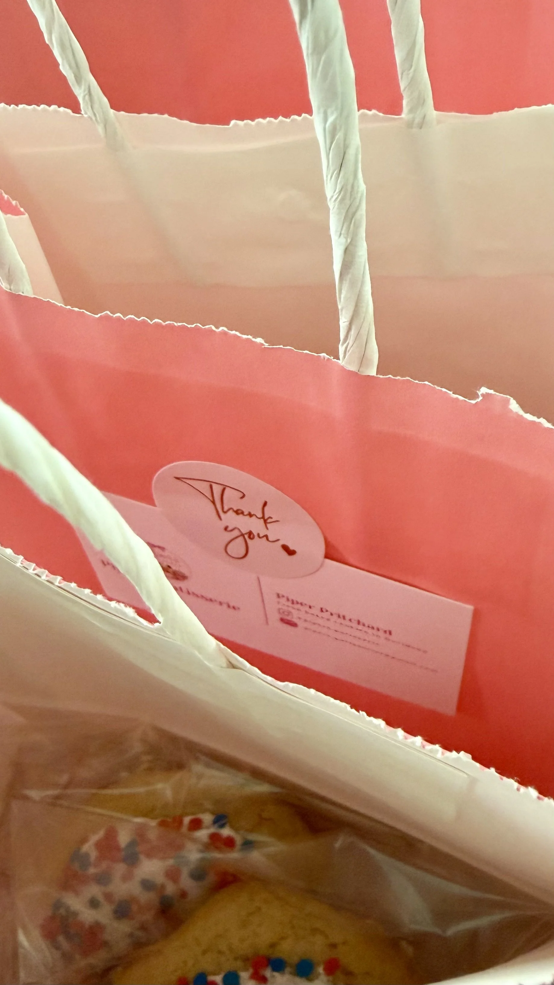 Inside a gift bag with pink and cream-colored tissue paper, a thank you card, and some small colorful items at the bottom.