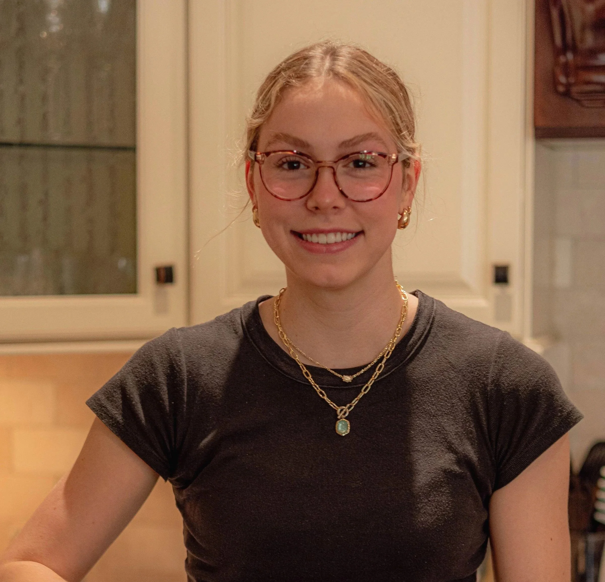 Close-up of a woman with blonde hair, wearing glasses, gold jewelry, and a dark gray top, smiling in a cozy indoor setting.