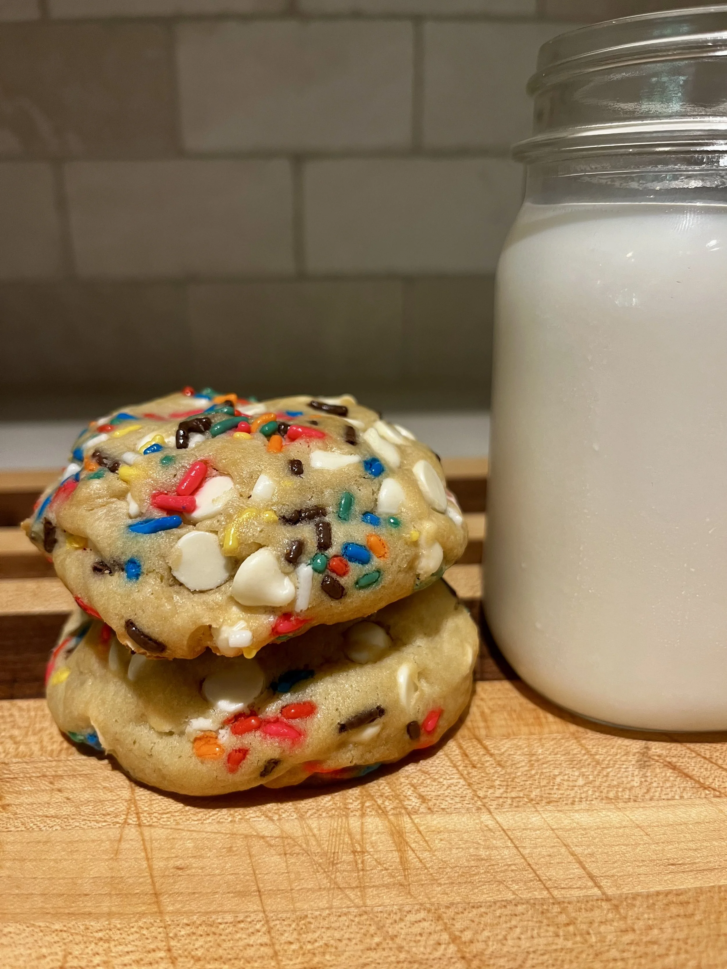 Close-up of two cookies with sprinkles and a glass jar of milk on a wooden surface.