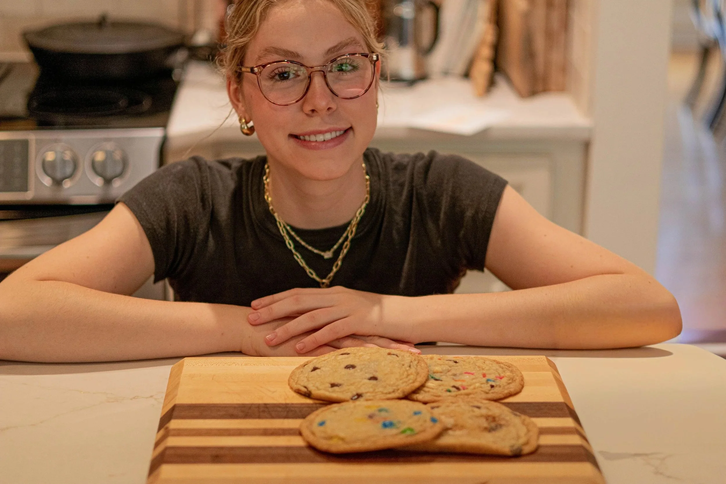 Portrait of a smiling woman with curly blonde hair and glasses, sitting at a table with a plate of cookies in front of her in a cozy kitchen.