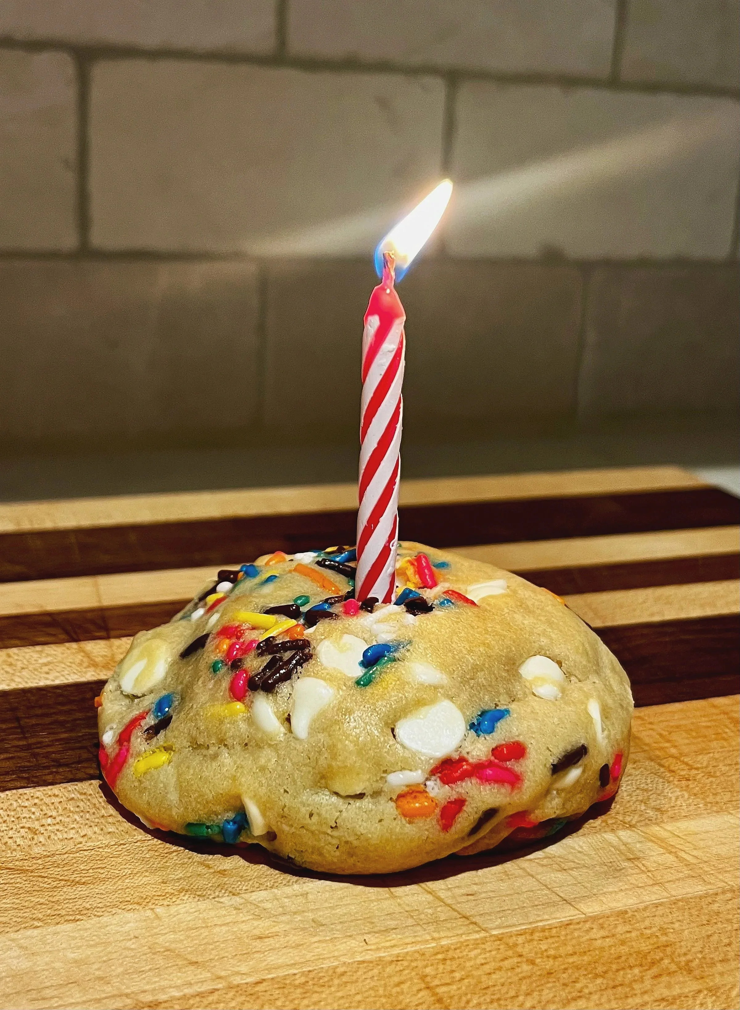 A close-up of a sugar cookie with colorful sprinkles, topped with a lit birthday candle, on a wooden surface.