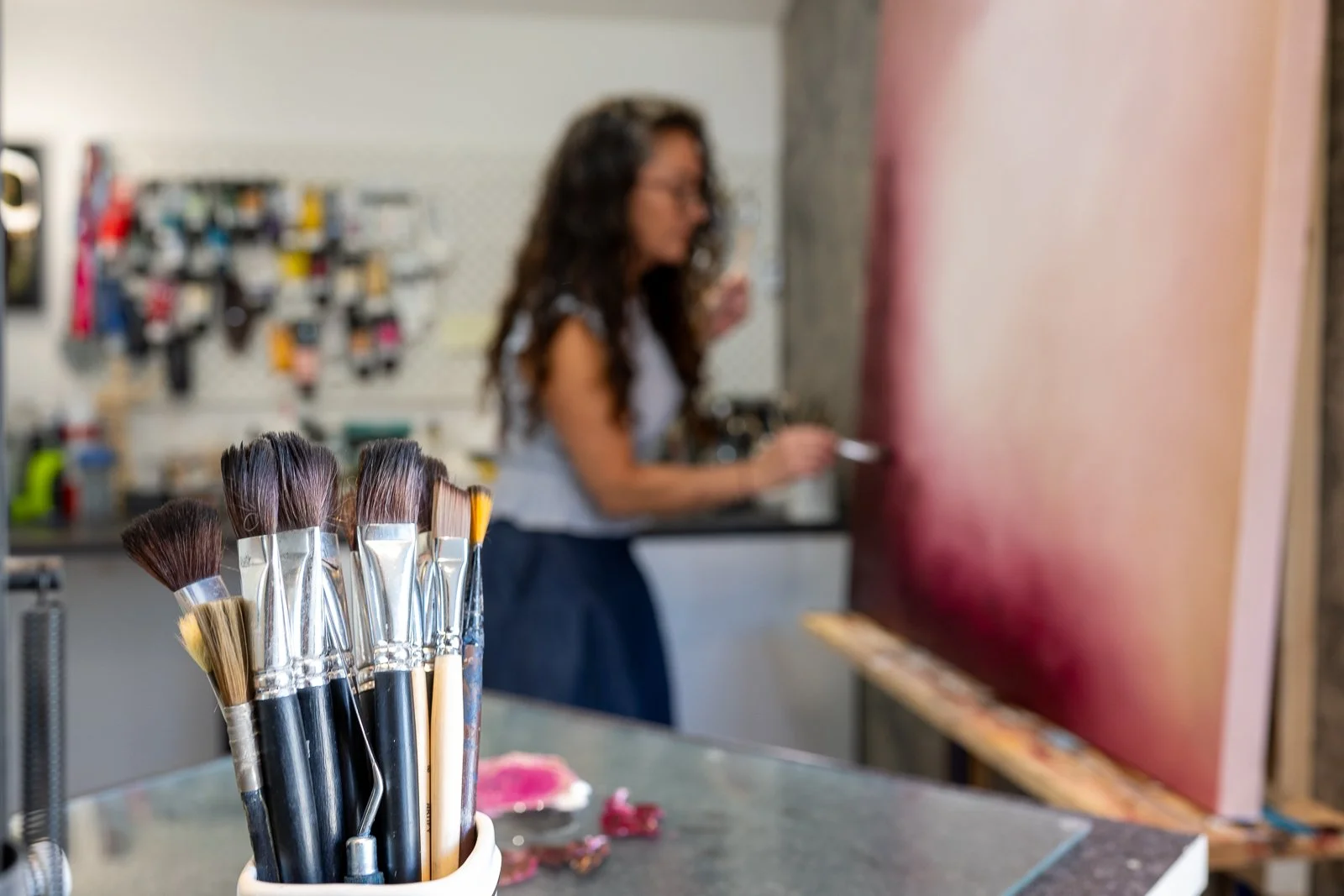 A collection of paintbrushes in the foreground with a woman painting on a canvas in the background.