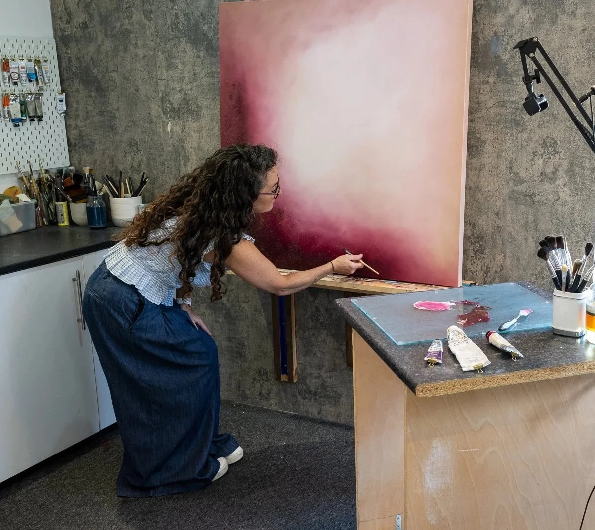 A woman with curly hair and glasses is painting an abstract sunset scene on a large canvas in an art studio. She is leaning forward, using a brush to blend colors of pink, orange, and white. The studio has a concrete wall, a pegboard with art supplies, and a countertop with paint tubes and brushes. A lamp arm is visible in the upper right corner.