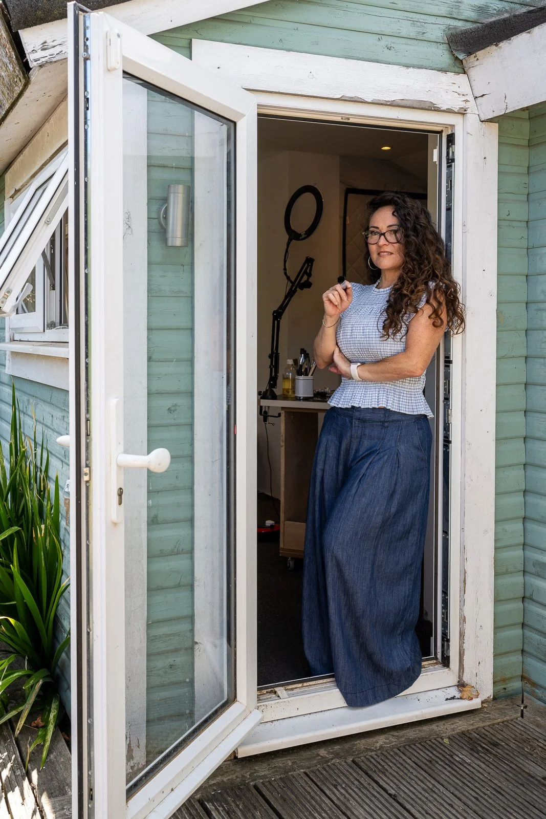 A woman with curly hair, glasses, and wearing a blue and white checkered top with wide-leg jeans stands at a door, holding a small object, with a ring light and recording equipment inside.