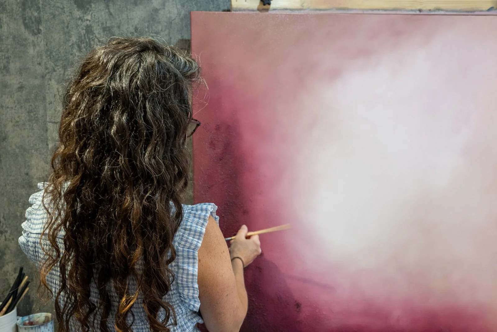 Woman with long, curly hair painting a pink and white wall with a brush.