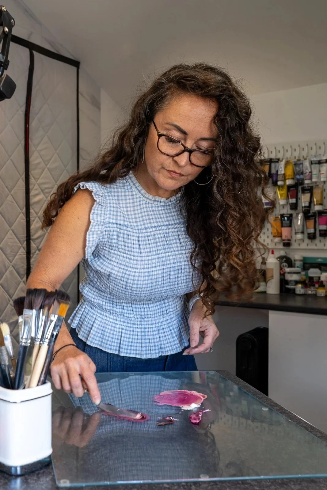 A woman with long curly hair, wearing glasses and a blue checked sleeveless top, is standing at a table with some pink paint or pigment on it in a studio or workshop. There are paint brushes in a container on the table and art supplies on a wall in the background.