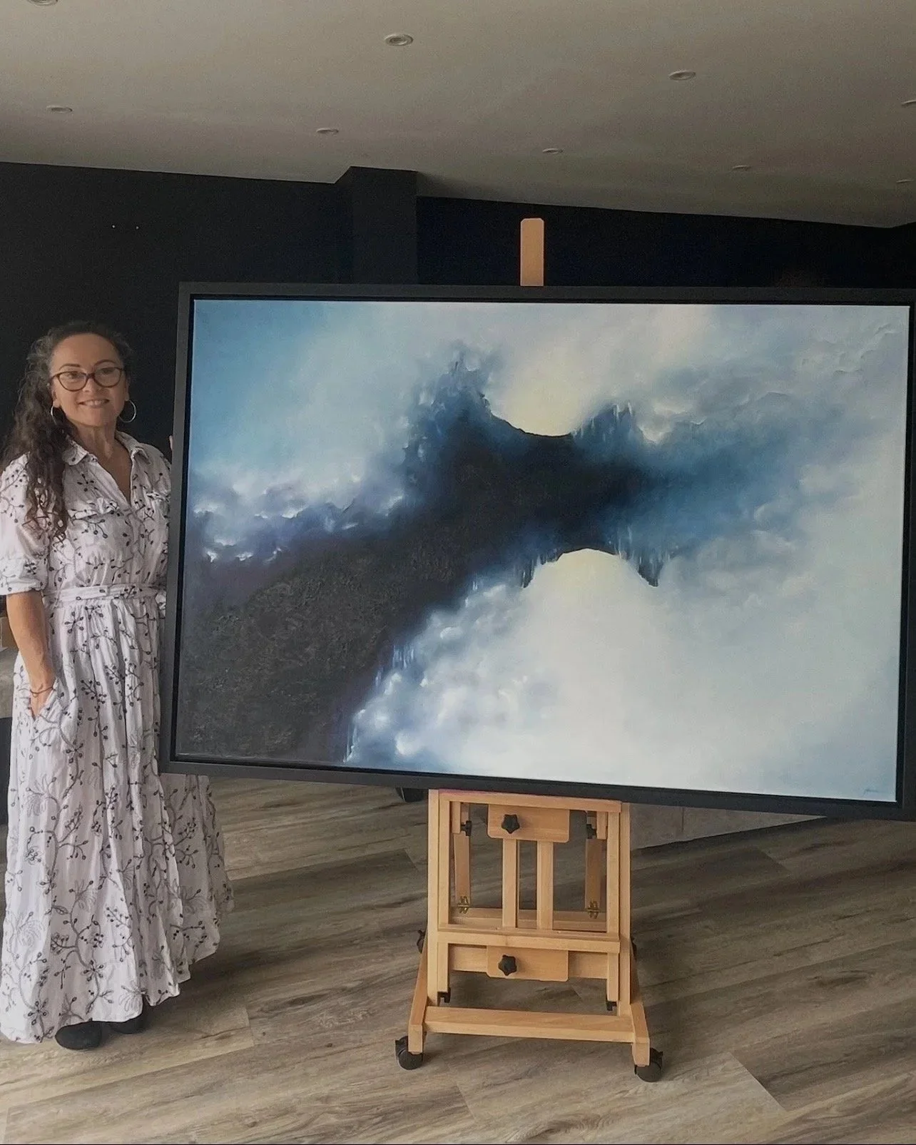 Mechelle Tebbs standing next to The Quiet Between Storms, abstract oil painting by Mechelle Tebbs,featuring deep blue cloud-like forms surrounding a passage of soft light.