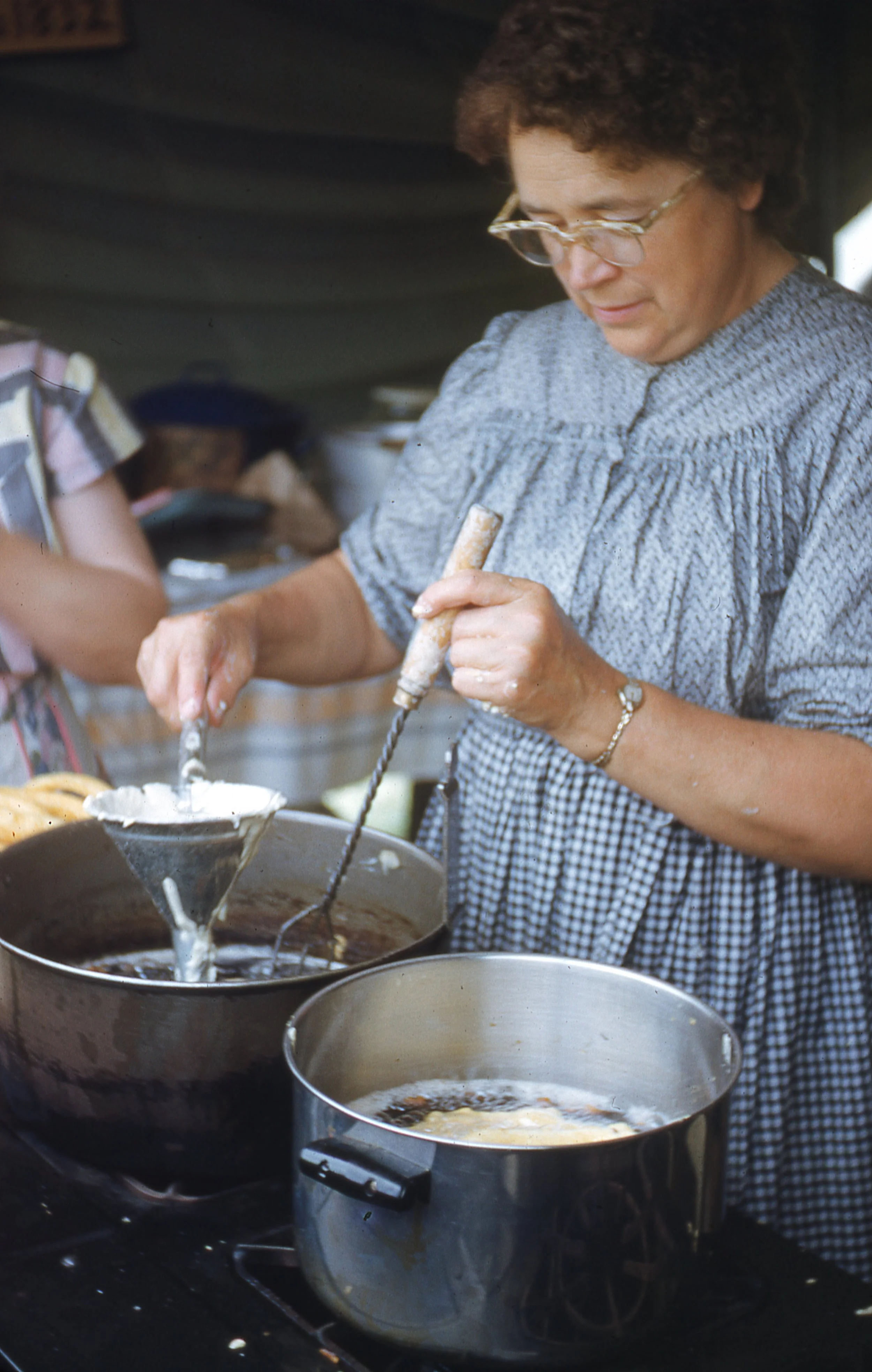 Woman, wearing long dress and apron, frying funnel cake in metal pot.