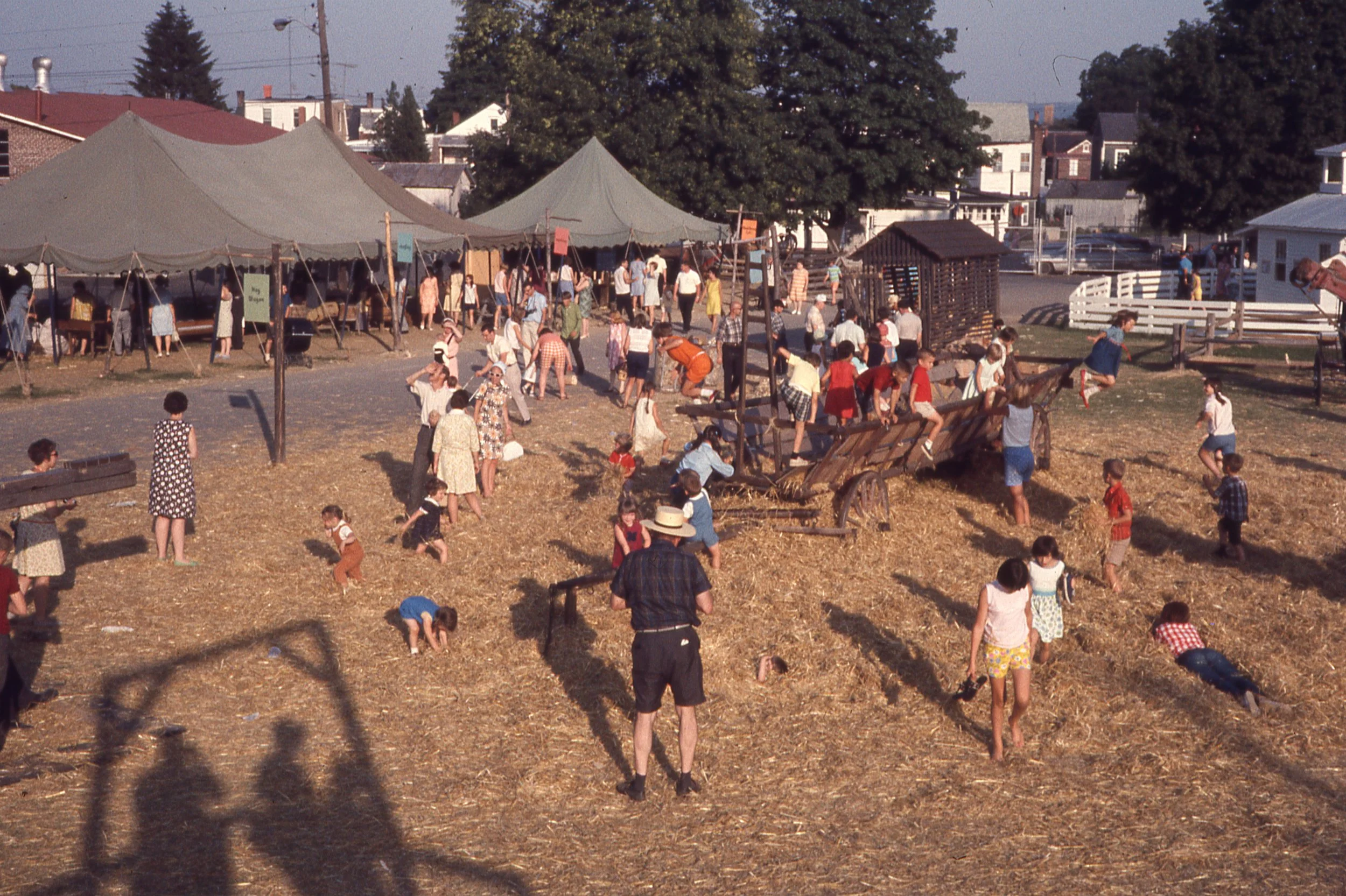 Large group of children and adults playing in straw and climbing on wooden wagon frame.