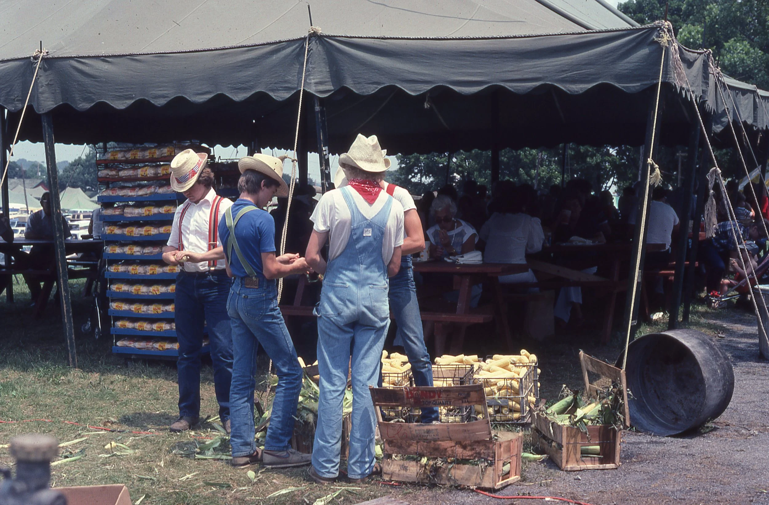 Group of young men in straw hats shucking corn beside food tent on Festival grounds.