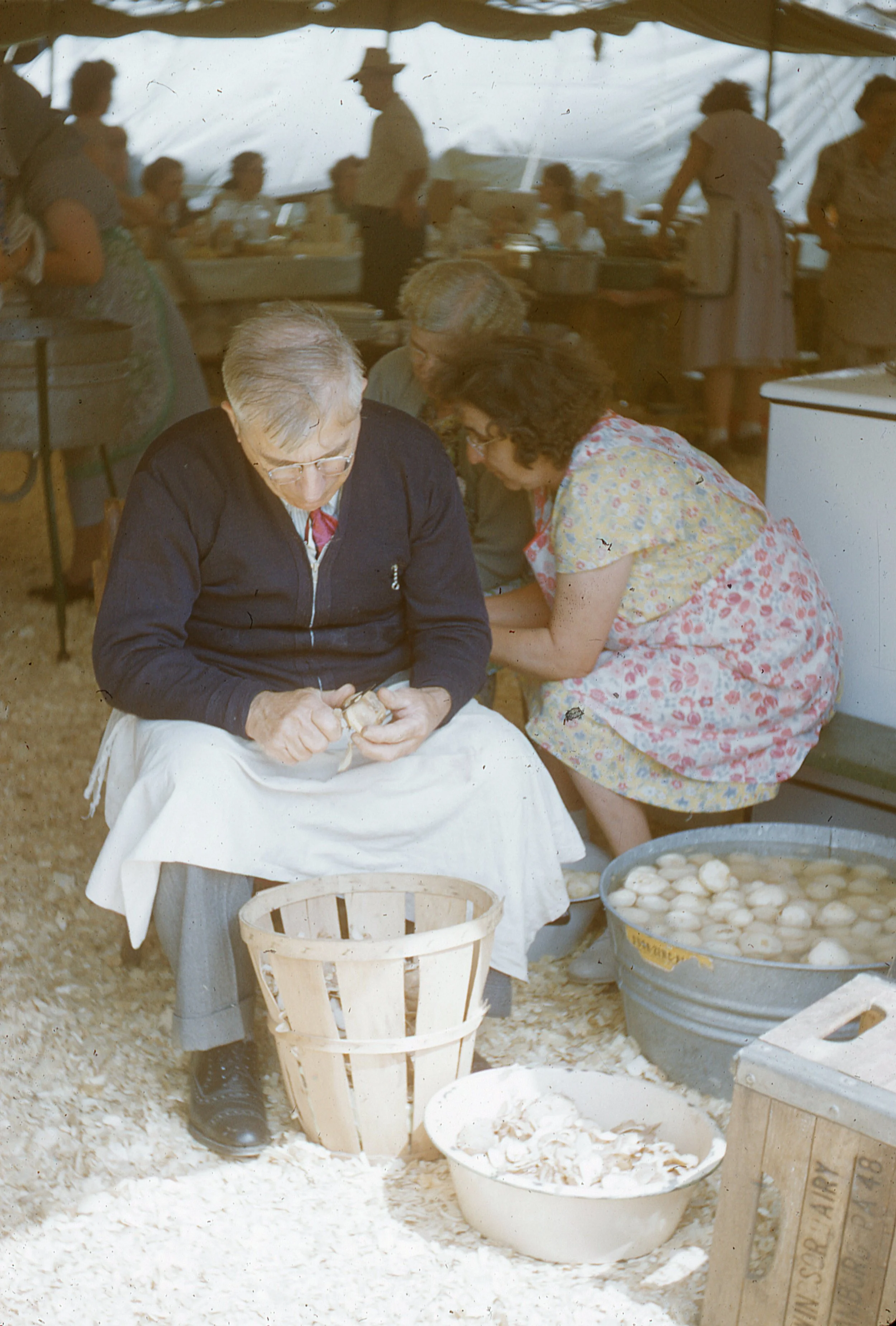 Man and two women peeling potatoes inside tent.