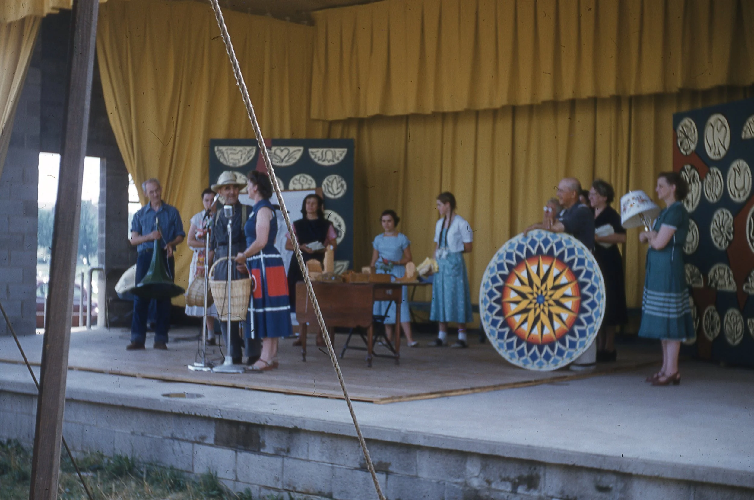 Man and women, both holding woven baskets, speaking on covered outdoor stage. Other presenters and crafts visible onstage behind speakers.