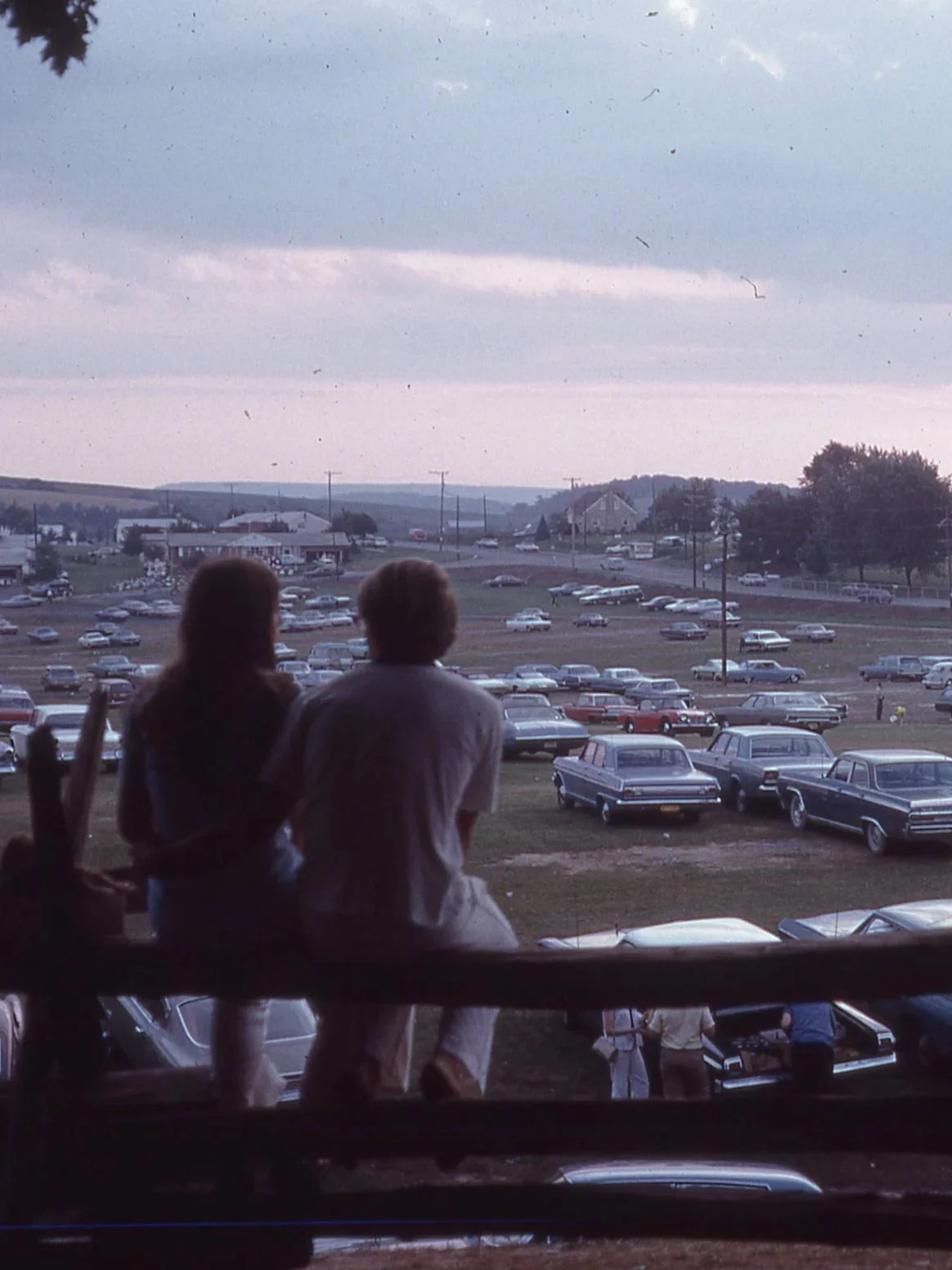Two people sitting on a wooden fence overlooking large, sparsely-filled parking area.