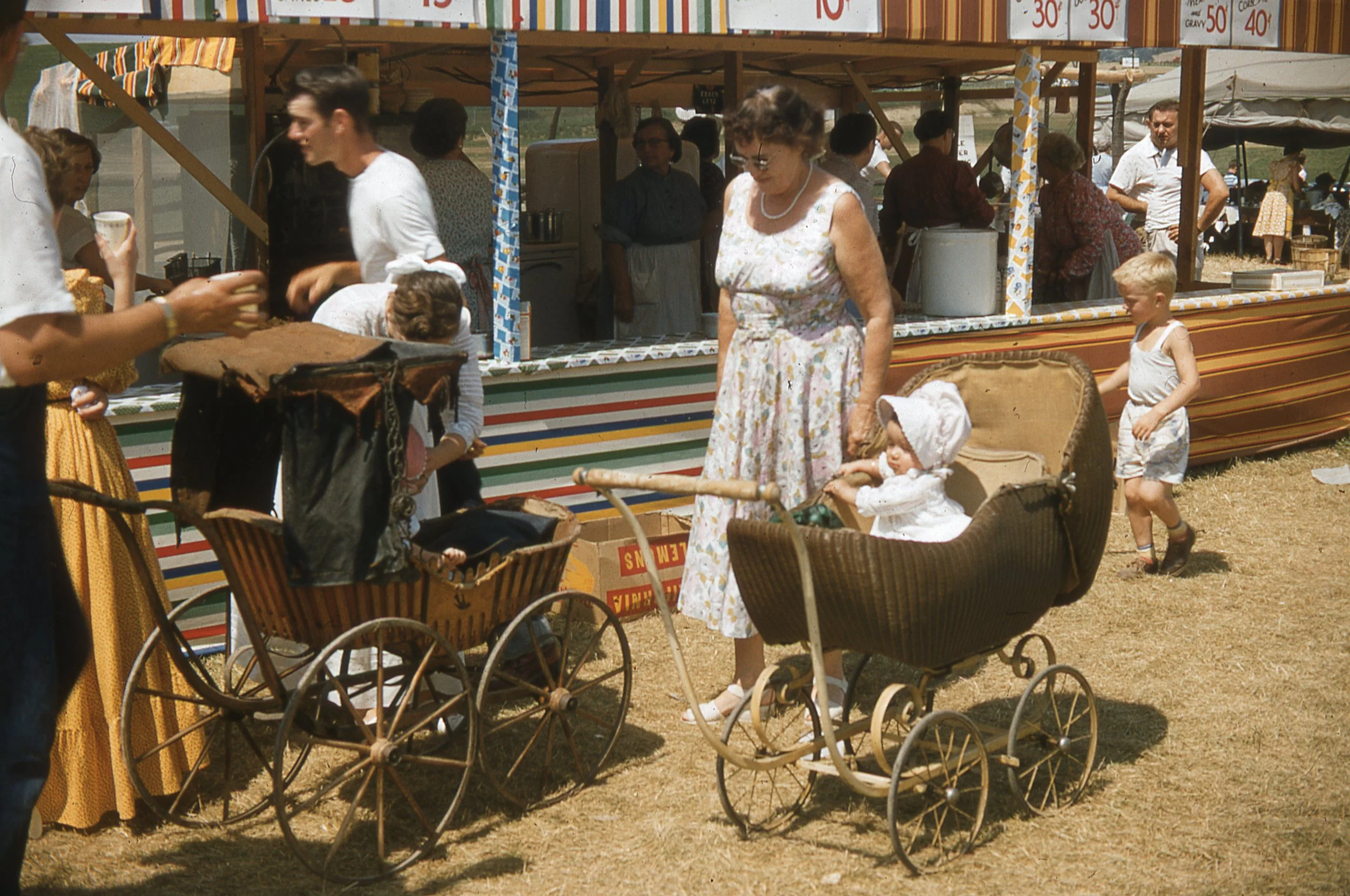 Two babies, in bonnets and white dresses, sitting in antique strollers in front of food stands.