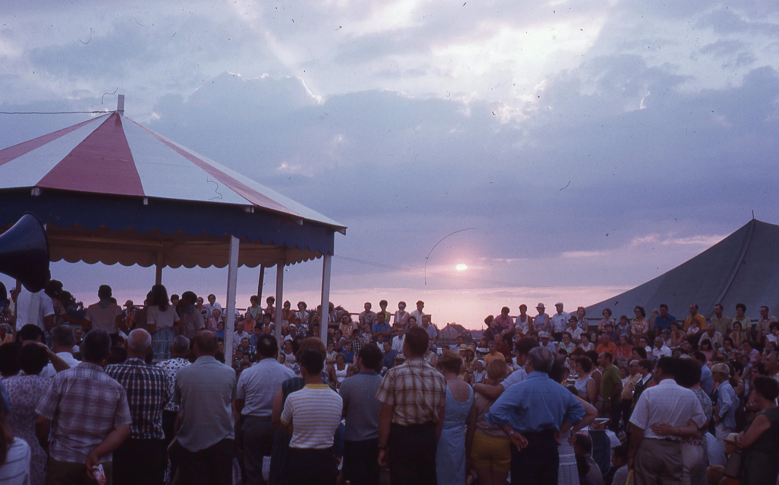 Large crowd watching performance on small outdoor stage at sunset.