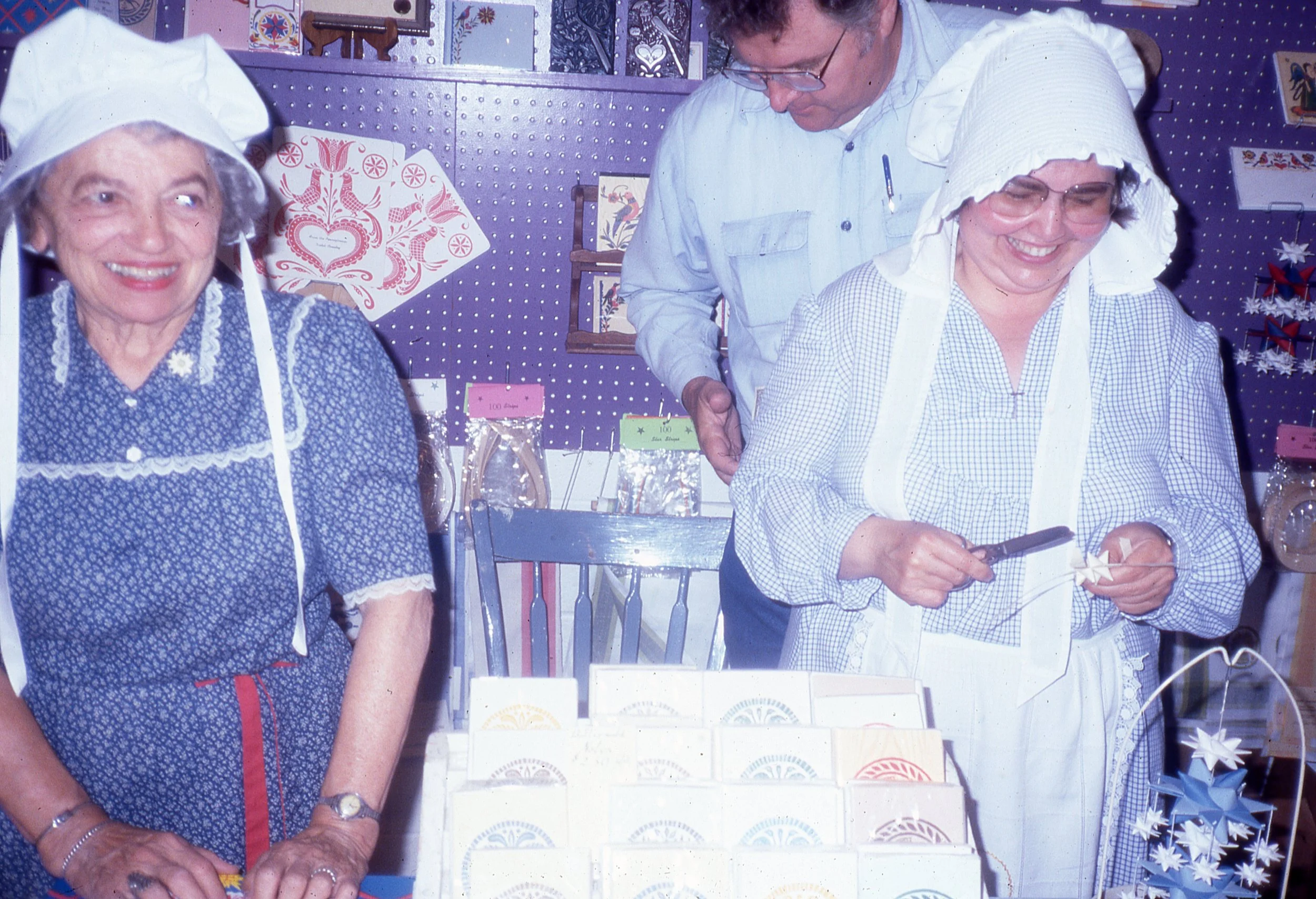 Two women, in aprons and bonnets, standing with man among display of paper star and printed paper cards and decoration.